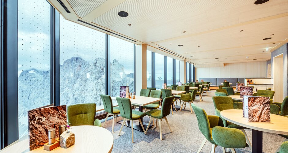 Helles Restaurant mit großen Panoramafenstern, grünen Polsterstühlen und Holztischen mit Blick auf die umliegenden Berge. | © Christoph Huber