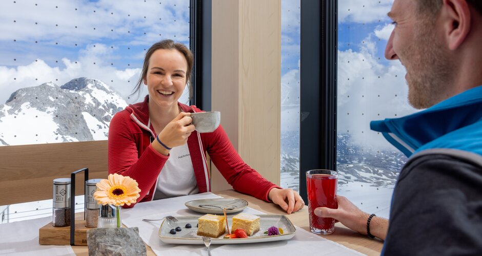 Lachende Frau in roter Jacke trinkt Kaffee, Mann mit rotem Getränk, Alpenpanorama durch große Fenster, Dessert auf Tisch. | © Harald Steiner