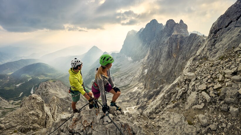 Two climbers with helmets and gear on a rocky ridge, gazing at the jagged Dachstein peaks under a dramatic sunset sky. | © Peter Burgstaller
