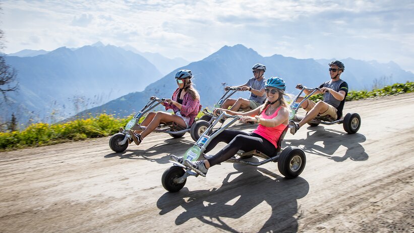 Vier Personen fahren mit Mountain-GoKarts bergab auf Schotterstraße mit Alpenblick und Sommerstimmung. | © Harald Steiner