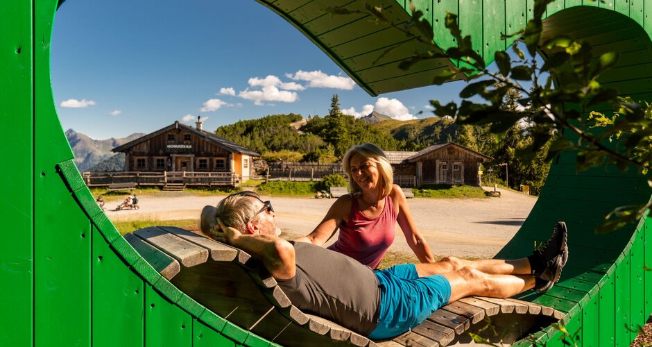 Wanderpaar entspannt auf Herzbankerl aus Holz mit Blick auf Almhütten und sonnige Berglandschaft. | © Christine Höflehner