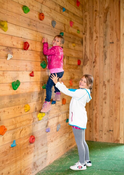 Mädchen hilft jüngerer Schwester beim Klettern an bunter Kletterwand aus Holz in Indoor-Spielbereich. | © Elisabeth Wolkenstein