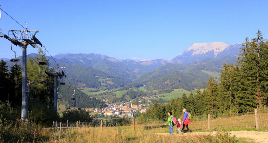 Family hikes along a meadow path with view of mountains, forests, chairlift and the village in the valley. | © Katharina Wassler