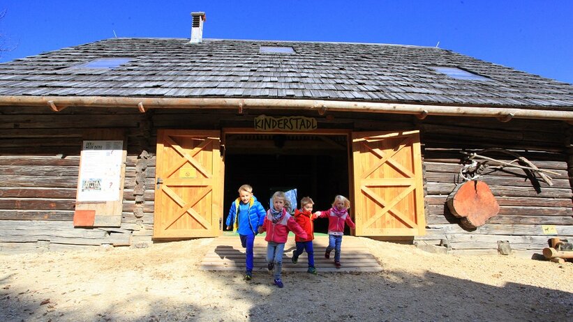 Five children run joyfully out of the wooden "Kinderstadl" building on a sunny day. | © Katharina Wassler