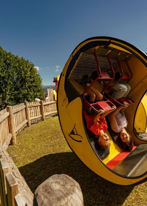 Two kids upside down in the yellow Moon Swing at Wilde Berg in Mautern, wooden fence and trees in background. | © Christine Höflehner