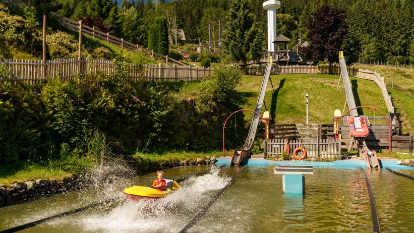 Child in yellow boat splashes into water at Der Wilde Berg in Mautern, wooden slides in the background. | © Christine Höflehner