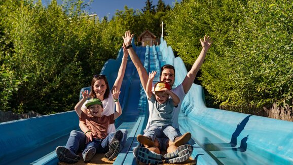 Two adults and two kids slide down a blue wave slide side by side, all laughing and raising their hands. | © Christine Höflehner