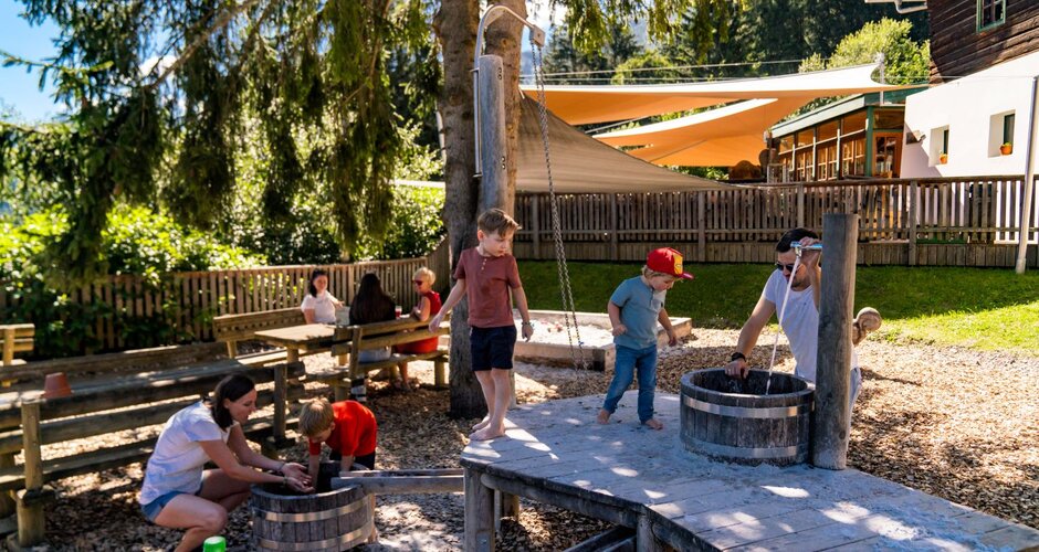 Children play barefoot with water and wooden barrels in the shaded playground area at Wilde Berg in Mautern. | © Christine Höflehner