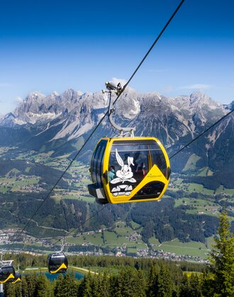 Yellow gondola with Hopsiland bunny above Planai with Dachstein mountains and green Enns valley in bright summer weather. | © Josh Absenger