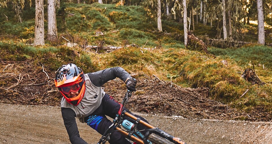 A mountain biker in helmet and goggles carves a sharp bermed turn on a forest trail in the bike park during golden sunlight. | © Armin Walcher
