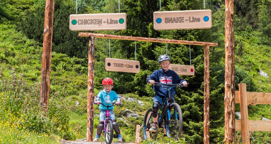 Two kids ride their mountain bikes into signed Junior Trails at Reiteralm, surrounded by wildflowers and green forest trees. | © Reiteralm Bergbahnen