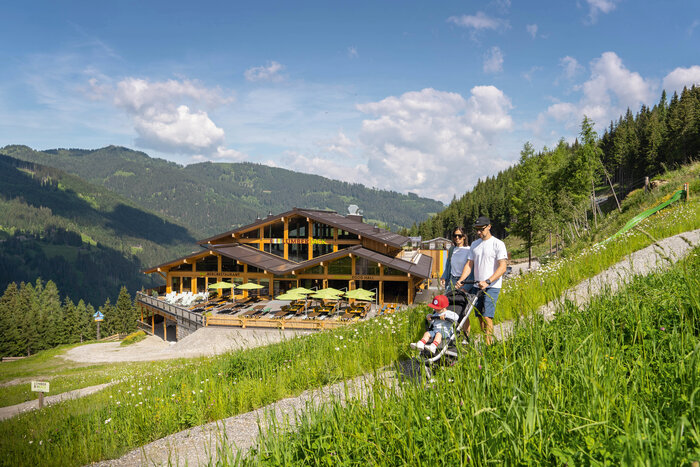 Familie mit Kinderwagen spaziert vor dem Bergrestaurant Lumberjack in grüner Berglandschaft. | © Shuttleberg