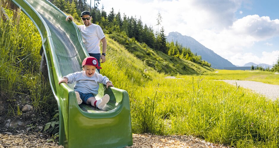 Ein Kleinkind rutscht lachend eine grüne Rutsche hinunter, dahinter steht ein Erwachsener. | © Shuttleberg