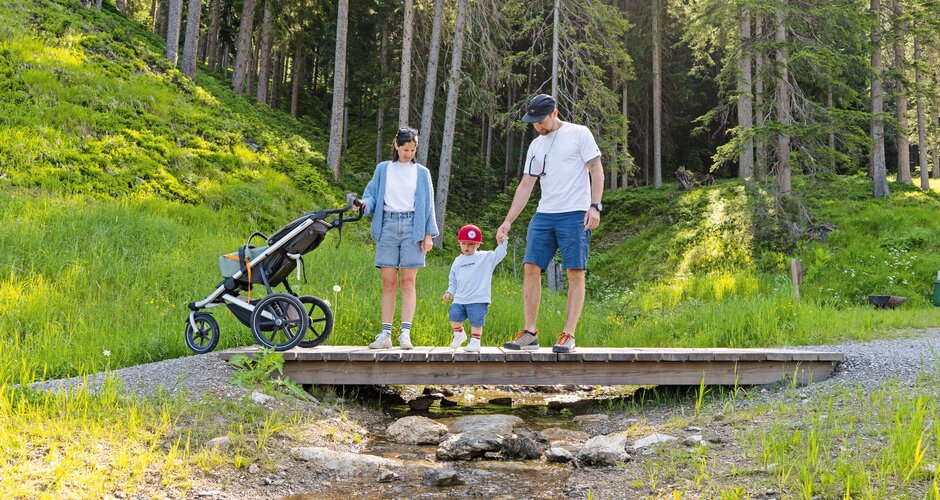 Eltern mit Kleinkind und Kinderwagen überqueren eine Holzbrücke im Grünen. | © Shuttleberg