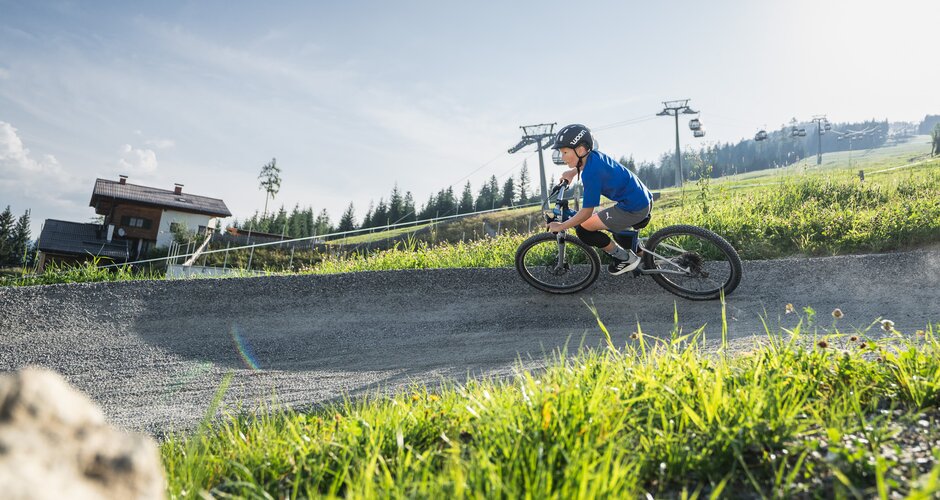 A boy in helmet rides a Woom bike on a curved trail with an alpine house, grassy slope, and gondola lift in the background. | © Snow Space Salzburg - Chris Eder