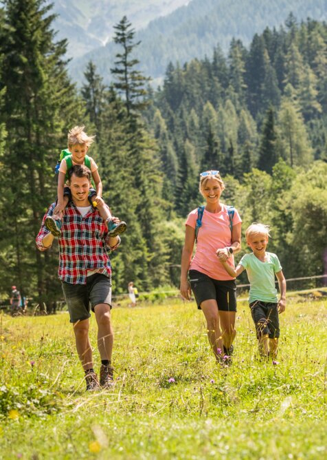Fröhliche Familie wandert über blühende Almwiese am Waldrand im Sonnenschein bei Sommerurlaub in den Alpen.
