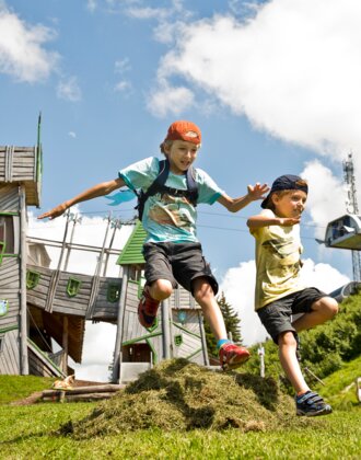 Two kids jump over a grassy mound in front of a fantasy play tower at Geisterberg adventure park in St. Johann, Salzburg | © sanktjohann.com | Mirja Geh