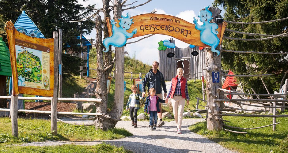 Family walking through signposted entrance to Ghost Village, colorful huts and overview map visible in the background | © Snow Space Salzburg