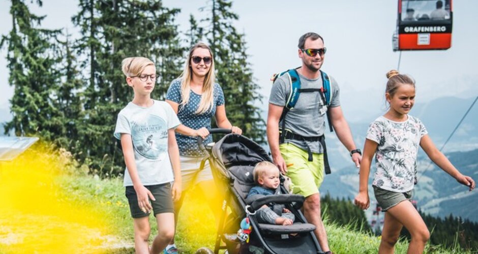 Family with stroller hiking along Grafenberg trail, a red gondola passes by in the background of the scenic view | © Snow Space Salzburg