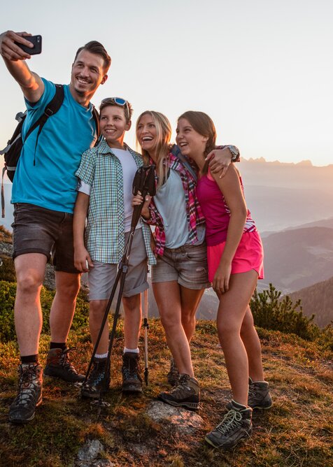 Family of four taking a selfie while hiking, sunset behind silhouettes of mountains in the scenic background