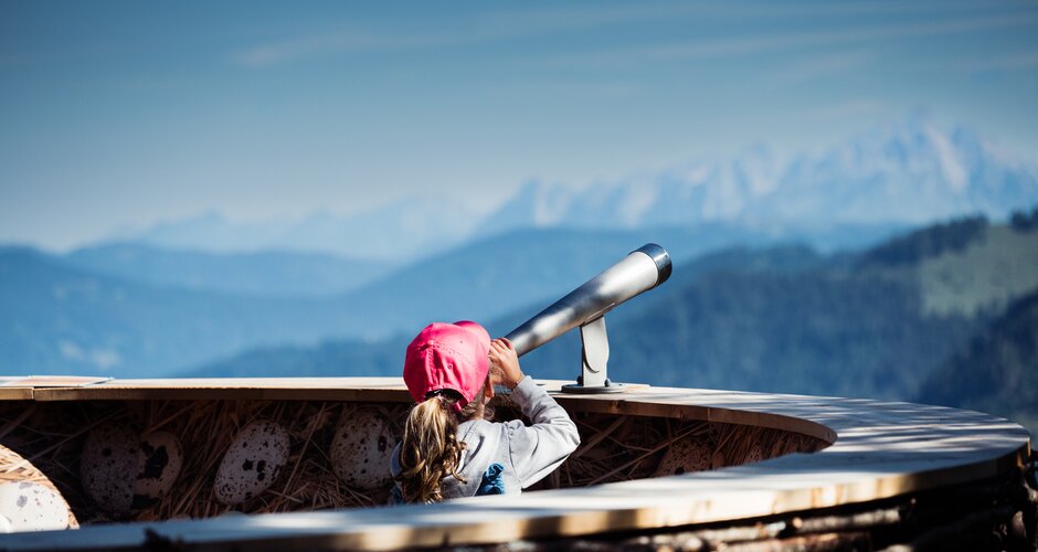 Mädchen mit roter Kappe blickt durch ein Fernrohr auf Bergpanorama von einer Aussichtsplattform in Zauchensee. | © Matthias Fritzenwallner