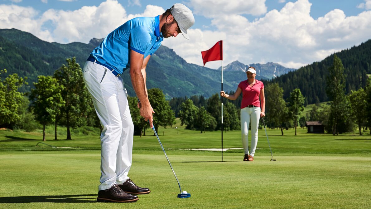 Man putting on a golf green, woman holding flag, mountains and trees in the background under a partly cloudy sky. | © TVB Radstadt_Paul Servern