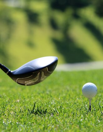 A golf ball on a tee about to be hit with a driver club, surrounded by bright green grass on a sunny fairway.