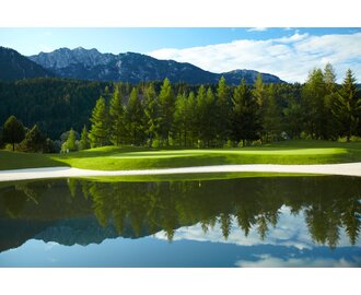 Grünes Fairway mit Fahne am Teichrand, gesäumt von Bäumen, mit Bergkulisse des Dachsteins im Hintergrund. | © GC Schladming-Dachstein