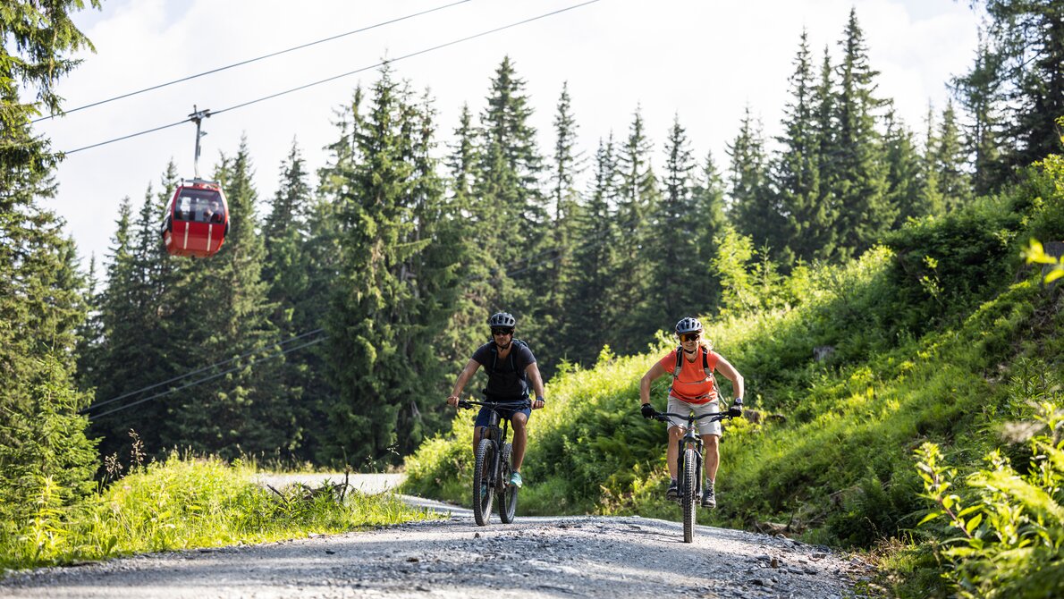 Two mountain bikers on gravel path below red gondola lift at Fulseck in lush green alpine scenery | © SalzburgerLand, Heiko Mandl