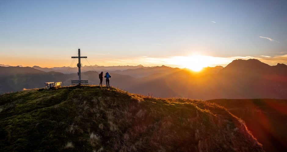 Two hikers at summit cross in Grossarl Valley during sunrise with wide mountain view and golden early morning light. | © Tourismusverband Großarltal 
