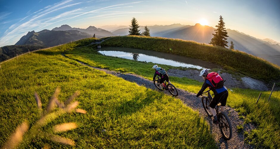 Two mountain bikers ride a narrow trail through green meadows at sunset with scenic views of the Grossarl Valley. | © mtb_travel