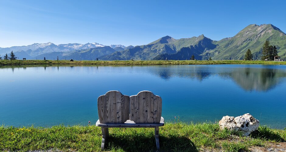 Wooden bench by Lake Himmelreich with calm water, mountain panorama and clear sky on a sunny summer day. | © TVB Großarltal