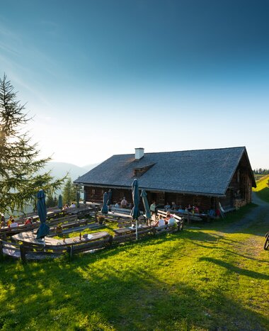 Evening atmosphere at Bichlalm with guests on terrace, bicycles in front and sun setting behind a larch tree. | © Gruber Michael