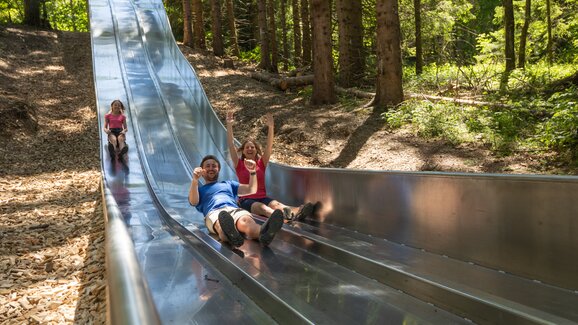 Family laughing while sliding down wide metal slide in forest slide park at Maria Alm's Natrun mountain | © Roland Haschka