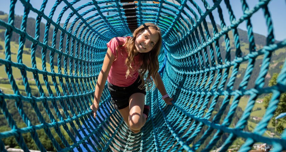 Smiling girl crawls through blue rope tunnel at Castle Playground Natrun, alpine meadow in background | © Roland Haschka