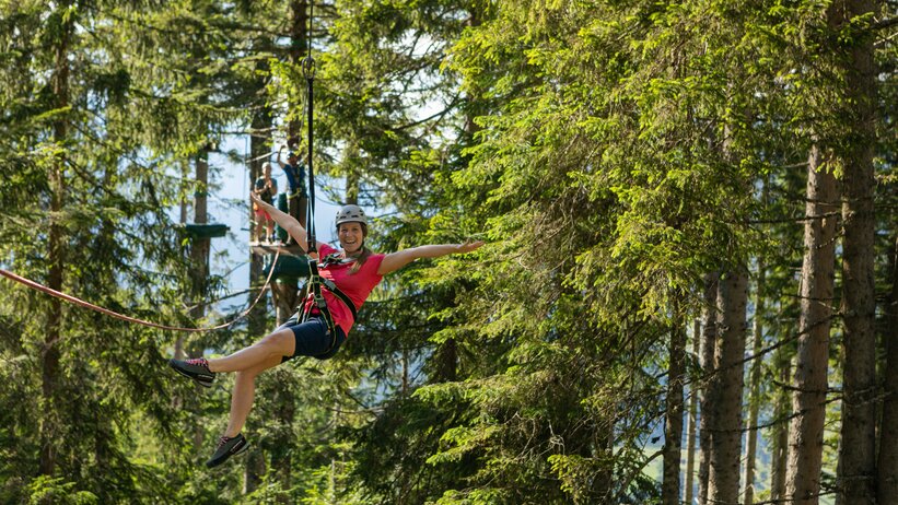 Smiling woman with helmet and harness rides zipline through forest rope park at Natrun in Maria Alm | © Roland Haschka