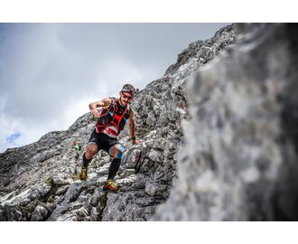 Sportler läuft bei Trailrunning-Rennen über felsigen Berghang, trägt Startnummer und Ausrüstung, weiterer Läufer im Hintergrund