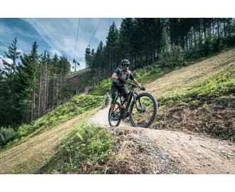 Biker mit Helm und Schutzkleidung fährt auf Flowtrail One in Maria Alm, im Hintergrund Gondelbahn und Wald | © Schartner