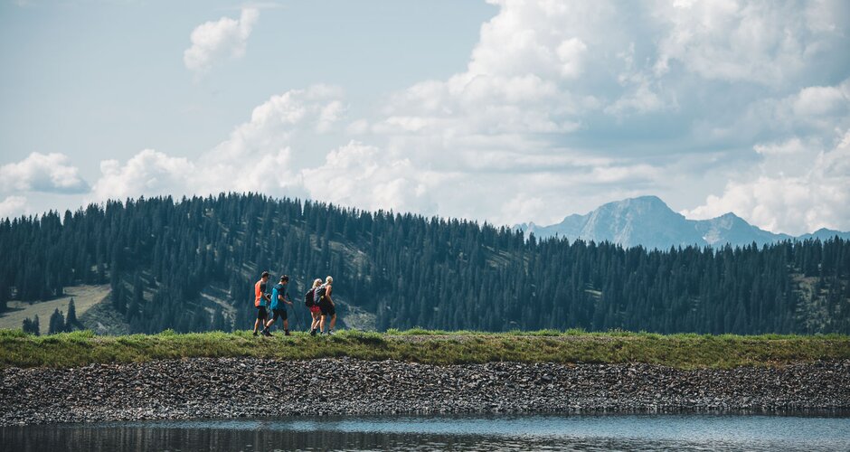 Four hikers walk along a mountain lake with forested hills and distant Hochkönig peaks under a partly cloudy summer sky | © Hochkönig Tourismus GmbH