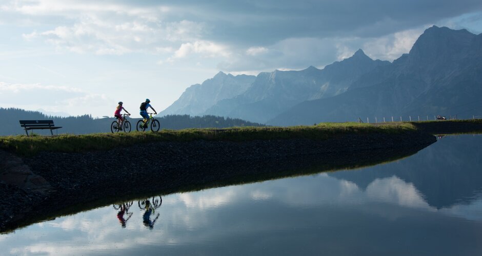 Two mountain bikers ride along calm reservoir, silhouette of Hochkönig mountains mirrored in the water | © Hochkönig Tourismus, Schartner