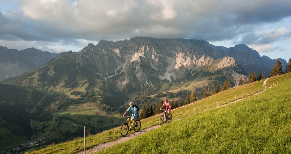Two mountain bikers ride alpine trail at sunset, rocky Hochkönig mountains glow in warm evening light | © Hochkönig Tourismus, Schartner