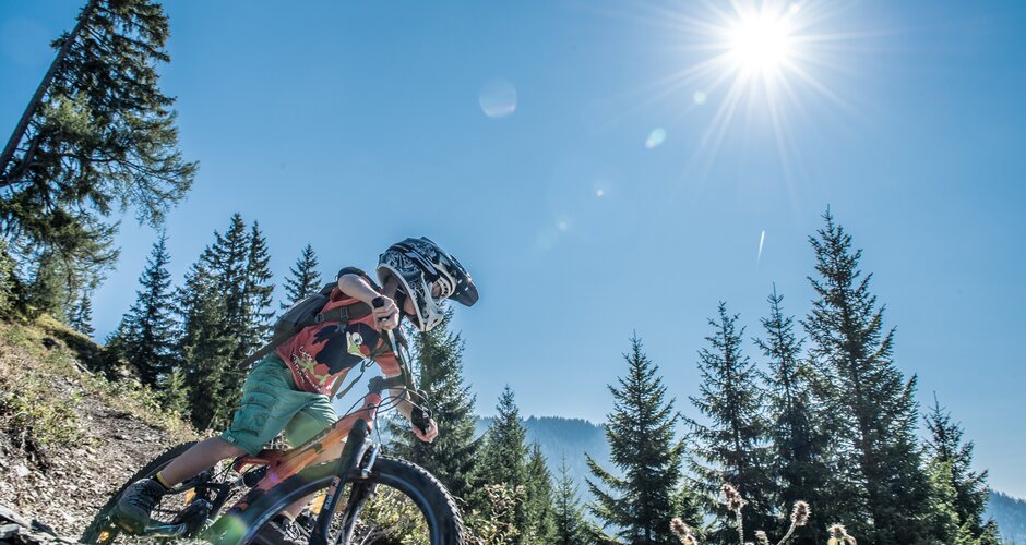 Kid with helmet biking a sunny Flowtrail in Dienten, pine forest and mountains in the background | © Hochkönig Tourismus, Creatina