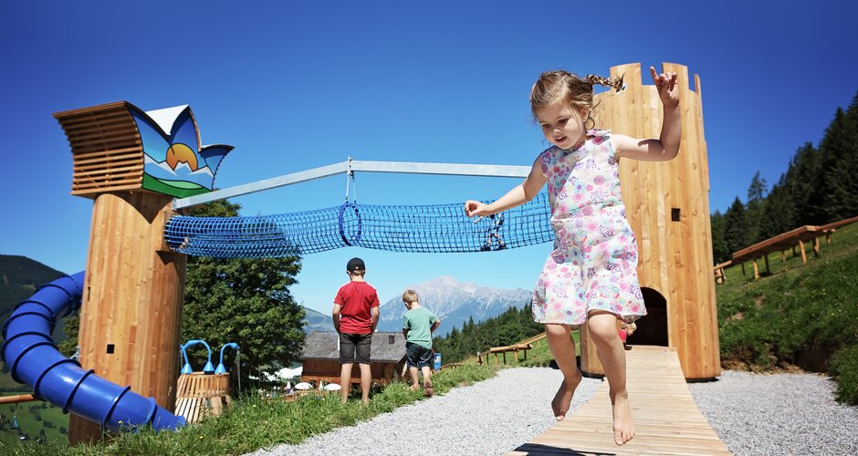 Barfüßiges Mädchen springt am Erlebnisspielplatz Natrun, im Hintergrund blaue Röhrenrutsche und Alpenpanorama