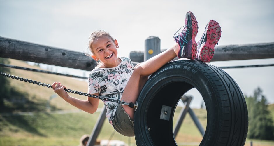 Smiling girl sits on a tire swing at the playground, holding onto chains, with grassy summer landscape in the background | © Salzburger Sportwelt