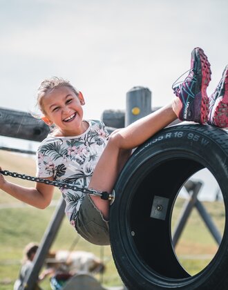 Smiling girl sits on a tire swing at the playground, holding onto chains, with grassy summer landscape in the background | © Salzburger Sportwelt