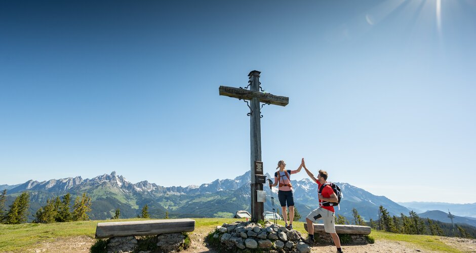 Two people stand on a plateau by a summit cross and give each other a high five | © Tourismusverband Radstadt/Lorenz Masser