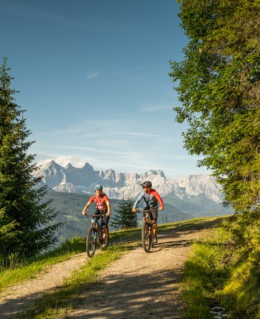 Two mountain bikers ride on a forest trail with a view of the Dachstein massif, surrounded by green trees and meadows. | © Altenmarkt-Zauchensee Tourismus/Lorenz Masser