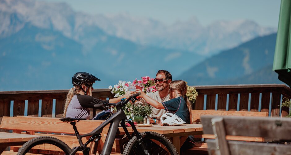 Family with e-bikes enjoys drink break at hut with mountain panorama in sunny weather | © O'MANS
