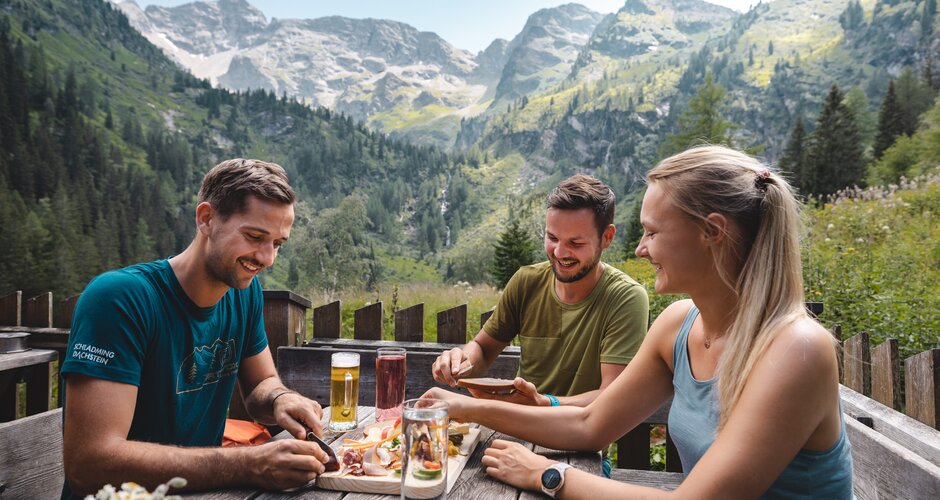 Three friends enjoy an alpine snack at a mountain hut in Seewigtal with forested slopes and high peaks in the background | © Mathäus Gartner