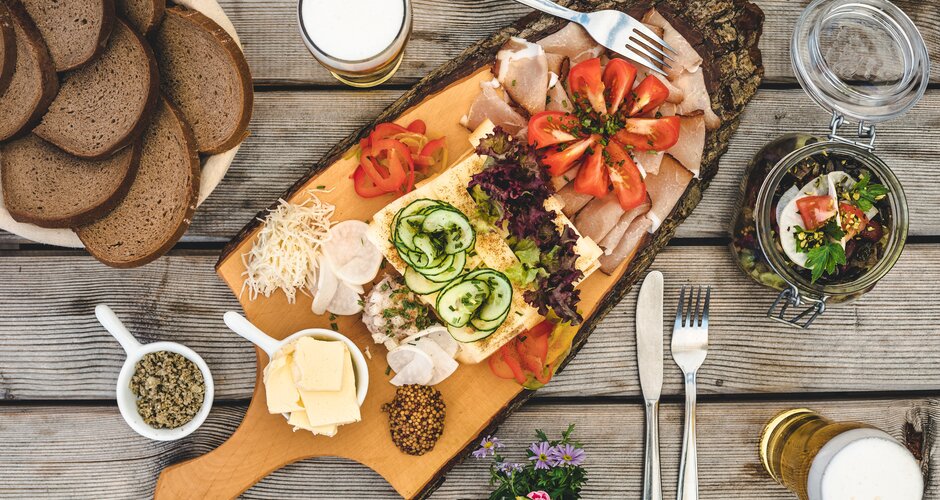 Hearty alpine snack with cheese, bacon, veggies, butter, bread and beer on wood board, plus salad in a glass jar | © Mathäus Gartner